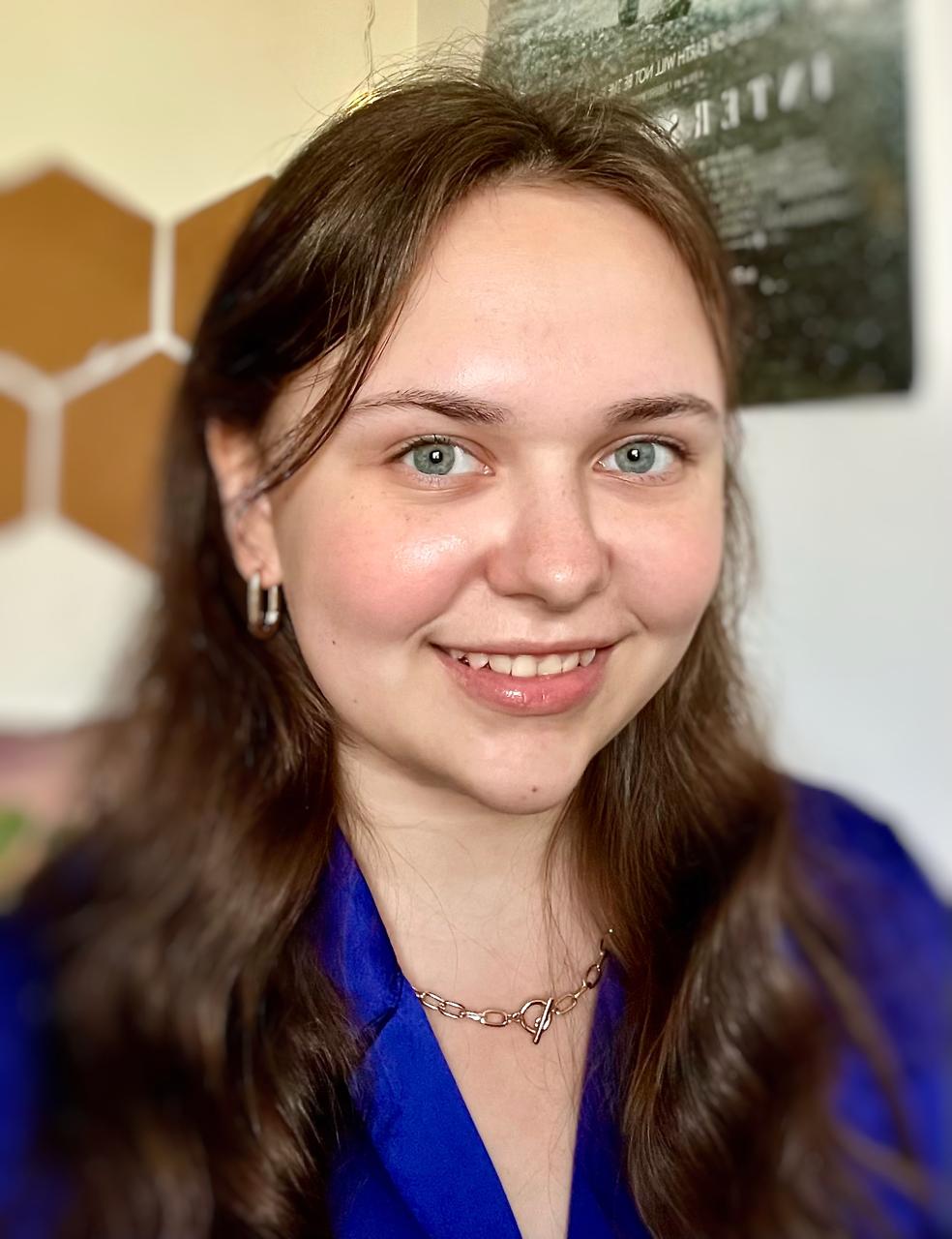 A smiling young woman looks at the camera. She is wearing dark blue clothing and a necklace.